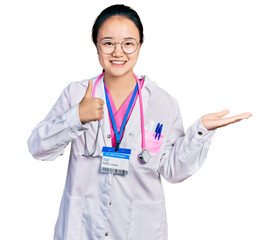 Young chinese woman wearing doctor uniform and stethoscope showing palm hand and doing ok gesture with thumbs up, smiling happy and cheerful