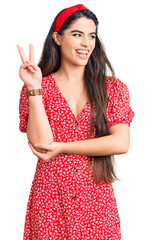 Brunette teenager girl wearing summer dress smiling with happy face winking at the camera doing victory sign with fingers. number two.