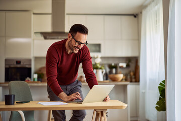 A happy adult male digital marketer leaning on a desk and checking his work on a laptop at home.