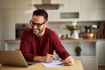 A smiling male freelancer with glasses writing notes on a paper while having an online meeting with a client on a laptop. - obrazy, fototapety, plakaty