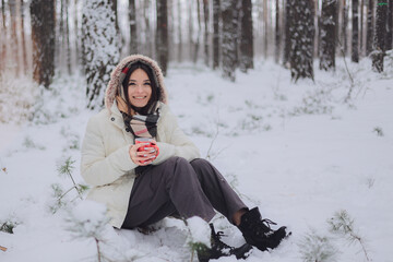 Portrait of a young pretty girl in a winter snowy forest. Happy girl in winter scenery, holding a red cup in her hand. Portret młodej, ładnej, uśmiechniętej  dziewczyny w zimowym,śnieżnym lesie.  - obrazy, fototapety, plakaty
