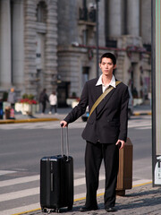 Portrait of handsome Chinese young man with black short hair wearing black blazer holding suitcase posing with modern city building background in sunny winter day, male fashion, cool Asian young man.