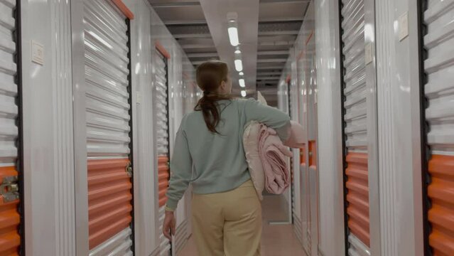 Rear view of woman with belongings enters in the self-storage facility unit. White and orange closed metal doors of storage warehouse. Moving, organizing, storage concept.