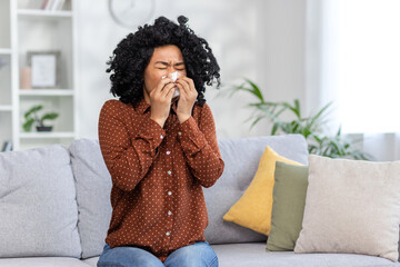 Sick young African-American woman sitting on sofa at home and wiping nose from runny nose with...