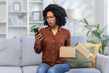 Worried young African American woman sitting on sofa at home, holding received open parcel in hand...