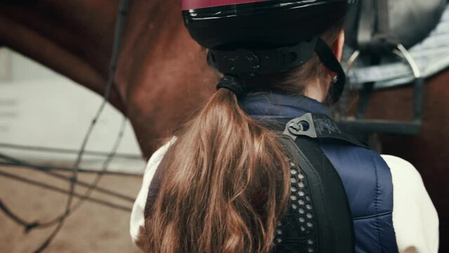 Little girl, kid in helmet taking care preparing to train. Child training horseback riding in special arena, pavilion. Concept of equestrian sports, childhood, school, course, active lifestyle, hobby