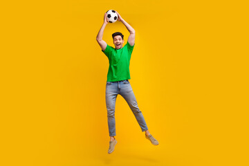 Fototapeta premium Full length photo of positive active man dressed green t-shirt jumping holding soccer ball over head isolated on yellow color background