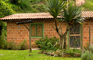 Casa com parede de tijolinho a vista, janela de madeira, jardim com planta e grama, sino dependurado em haste de madeira e serra com estrada ao fundo.