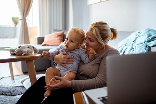 Mother Holding Toddler While Sitting On The Couch With A Laptop Nearby