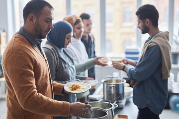 Side view portrait of adult Middle Eastern man receiving free hot meals at soup kitchen with people...