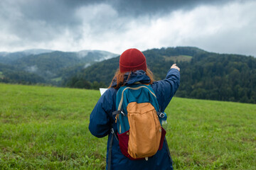 Stylish hipster 30s woman holding paper map, wearing backpack looking at mountain view while...