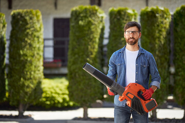 Strong, attractive man in safety glasses posing with modern cordless leaf blower outdoors. Portrait of smiling bearded gardener with powerful garden equipment, on blurred background. Concept of work.