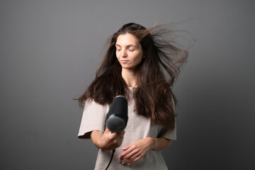 A content young woman is seen styling her flowing brown hair with a blow dryer against a neutral...