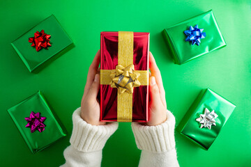 First person top view photo of hand holding red giftbox with yellow satin ribbon bow over mini colored presents on isolated green background with copyspace.