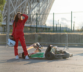 determined girl in a red suit holding a helmet  involved in kart racing stands next to a go-kart,...