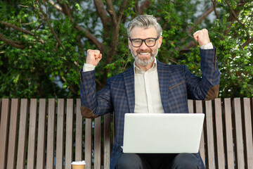 Joyful senior businessman outdoors raising his fists in victory, with laptop and coffee, showcasing success and achievement.