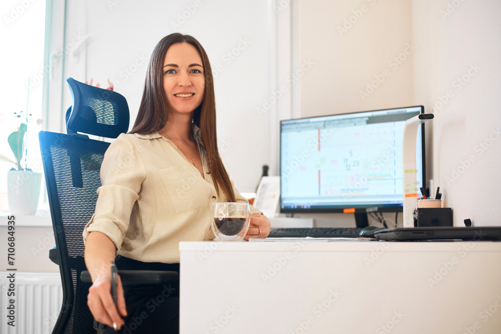 Wall mural Portrait of a happy female designer estimator sitting at the table with a notebook and calculator, counting the balance and typing on a laptop, finishing her work with a cup of coffee.