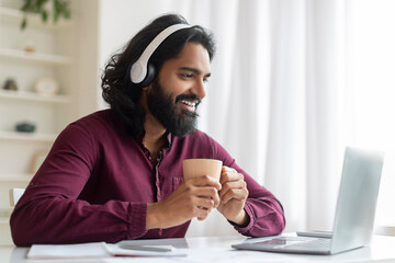 Joyful indian man with coffee and headphones working on his laptop