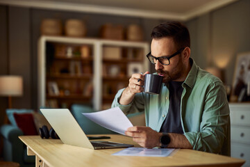 A male freelancer drinking coffee while analyzing his work on a laptop with a document in his hand.