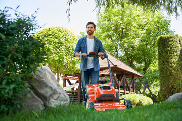 Smiling guy with beard trimming overgrown green lawn with electric mower in garden. Low angle view...