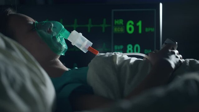 A female patient in medical mask is breathing oxygen support in a hospital next to heart rate machine. Unhealthy woman in coma. Getting mechanical ventilation. Concept of pandemic and therapy.