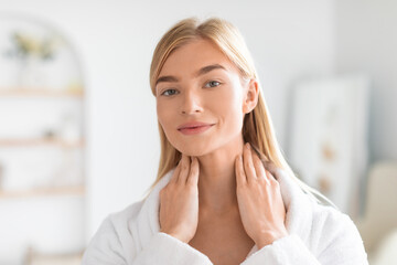 blonde woman in bathrobe massaging skincare lotion on neck indoor