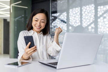 A cheerful young Asian businesswoman exudes success as she celebrates with a fist pump at her office workstation, laptop and phone in hand.