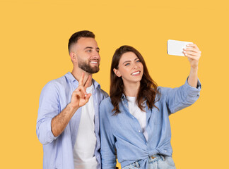 Happy couple taking a selfie, making peace sign on a yellow background