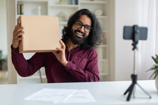 Happy Indian man presenting cardboard box to smartphone camera, recording unboxing video