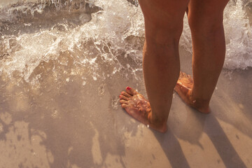 The naked feet of a woman standing on the beach next to the water, on a clear sand, a smooth wave...