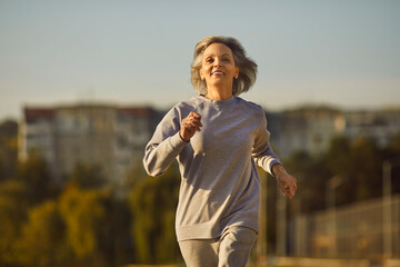Happy beautiful smiling senior woman in sports clothes running on a city street during her outdoor fitness workout in the summer. Old age, healthy lifestyle, exercise concept - obrazy, fototapety, plakaty