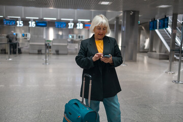 Smiling 60s senior business woman passenger in black jacket with suitcase using phone in terminal...
