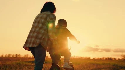 Foto op Plexiglas Mom teaches her son to ride bike in park at sunset. Family day out in nature. Mother little son learn to ride bike together in summer. Boy, his mother learn to ride bicycle in meadow in sun. Teamwork © zoteva87