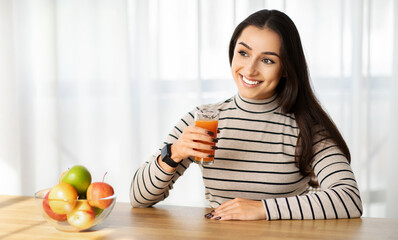 Cheerful young woman enjoying a fresh glass of juice, with a bowl of colorful fruits on the table