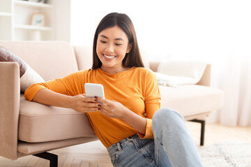 Cute young asian woman sitting on floor, using phone