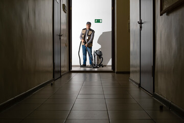 A man is vacuuming in a long hallway. The cleaner is at work.
