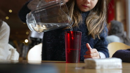 Child pouring water into red plastic cup with jar standing at restaurant, small girl serving refreshing tap water at diner