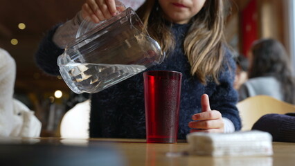 Child pouring water into red plastic cup with jar standing at restaurant, small girl serving...