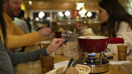 Closeup of Cheese Fondue Dining - Hand Dipping Bread in Melted Cheese, Red Pot Over Heater in Restaurant