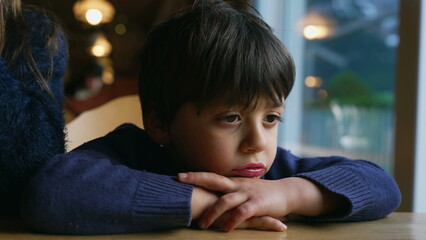 Pensive child feeling boredom at restaurant, leaning on table. Thoughtful little boy daydreaming,...