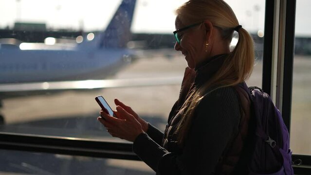 Mature woman traveler uses a smartphone in the airport terminal gets good news winner gesture. Airplane outside the window in the background.