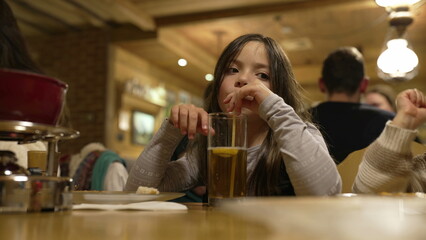 Little girl drinks ice tea with straw at restaurant in the evening. Child sipping drink at diner