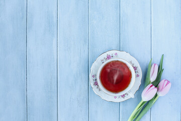 Steaming hot cup of tea and pink spring tulip flowers over a rustic blue wooden table. Table top view. Directly above.