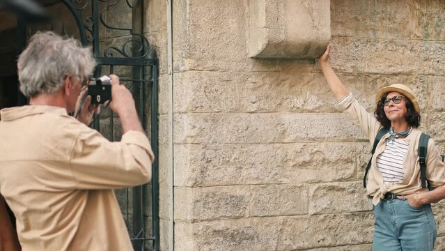 Camera View Of Older Caucasian Male Making Photo Of His Beloved Wife. Woman Leaning On Building And Posing For Best Photo. Man Trying To Catch Focus With Camera For Memorable Shot.