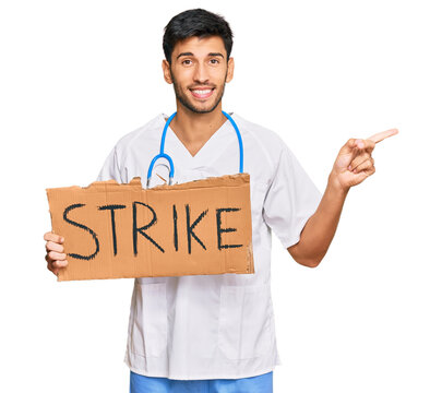 Young handsome man wearing doctor uniform holding strike banner cardboard smiling happy pointing with hand and finger to the side