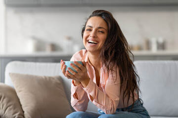 Joyful woman laughing while holding cup, in cozy home setting