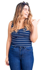 Young caucasian woman wearing casual clothes and sunglasses smiling with happy face looking and pointing to the side with thumb up.