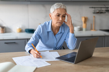 Stressed senior lady working on laptop taking notes touching head