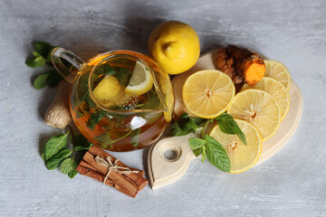 Glass teapot with ginger tea, lemon, turmeric root and mint on grey background with space for text. Cold and flu season concept. 