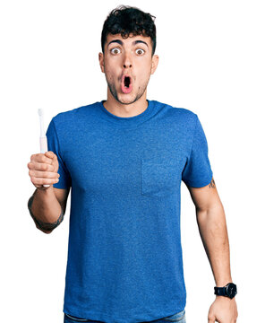 Young hispanic man holding electric toothbrush scared and amazed with open mouth for surprise, disbelief face